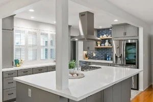 Gray kitchen with large island, cooktop, stainless hood, and navy tile backsplash with floating shelves.